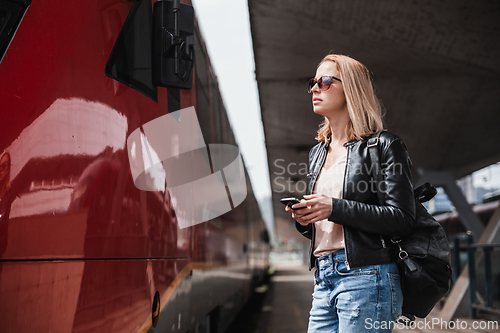 Image of Young blond woman in jeans, shirt and leather jacket wearing bag and sunglass, embarking red modern speed train on train station platform. Travel and transportation.