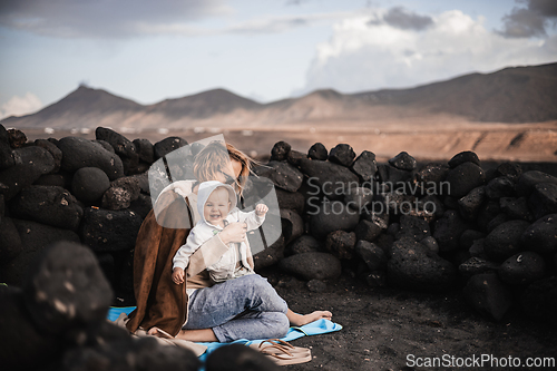 Image of Mother enjoying winter vacations playing with his infant baby boy son on black sandy volcanic beach of Janubio on Lanzarote island, Spain on windy overcast day. Family travel vacations concept.