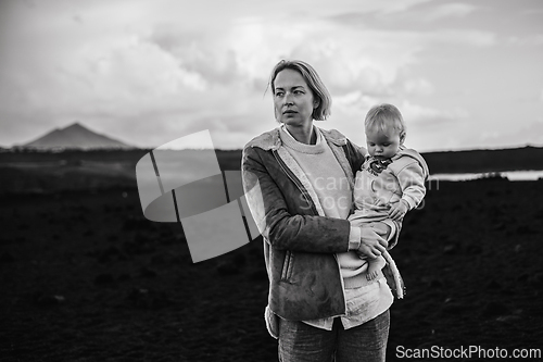 Image of Mother enjoying winter vacations playing with his infant baby boy son on black sandy volcanic beach of Janubio on Lanzarote island, Spain on windy overcast day. Family travel vacations concept.