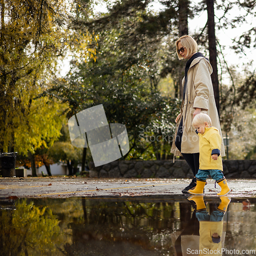 Image of Small bond infant boy wearing yellow rubber boots and yellow waterproof raincoat walking in puddles on a overcast rainy day holding her mother's hand. Mom with small child in rain outdoors.