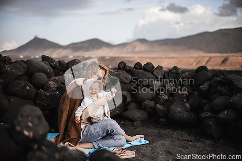 Image of Mother enjoying winter vacations playing with his infant baby boy son on black sandy volcanic beach of Janubio on Lanzarote island, Spain on windy overcast day. Family travel vacations concept.