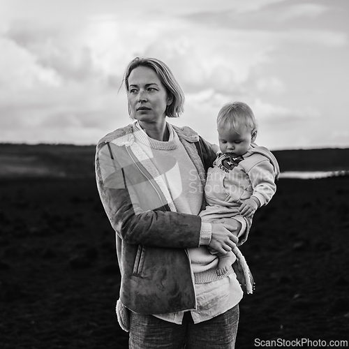 Image of Mother enjoying winter vacations playing with his infant baby boy son on black sandy volcanic beach of Janubio on Lanzarote island, Spain on windy overcast day. Family travel vacations concept.