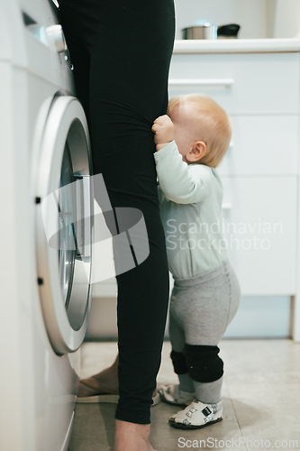 Image of Little infant baby boy child hiding between mothers legs demanding her attention while she is multitasking, trying to do some household chores in kitchen at home. Mother on maternity leave.