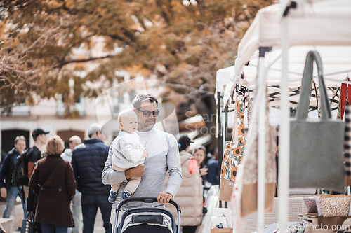 Image of Father walking carrying his infant baby boy child and pushing stroller in crowd of people visiting Sunday flea market in Malaga, Spain.
