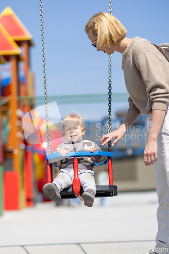 Image of Mother pushing her infant baby boy child on a swing on playground outdoors.