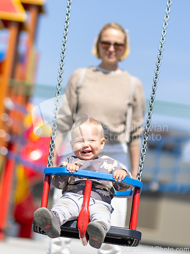 Image of Mother pushing her infant baby boy child on a swing on playground outdoors.