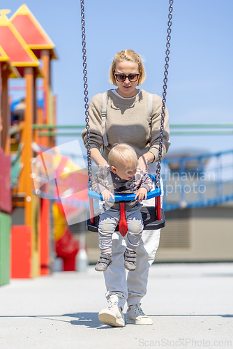Image of Mother pushing her infant baby boy child on a swing on playground outdoors.