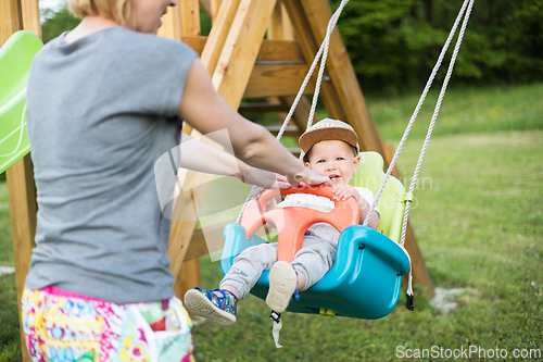 Image of Mother pushing her infant baby boy child on a swing on playground outdoors.