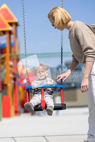 Image of Mother pushing her infant baby boy child on a swing on playground outdoors.