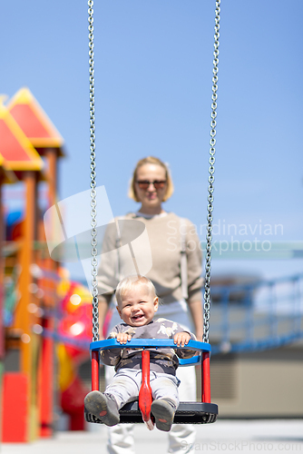 Image of Mother pushing her infant baby boy child on a swing on playground outdoors.