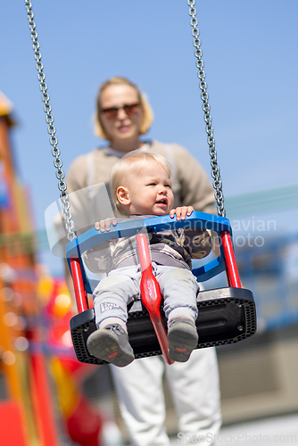 Image of Mother pushing her infant baby boy child on a swing on playground outdoors.