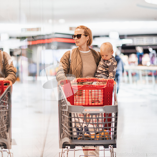Image of Mother shopping with his kid in the mall. Mother pushing shopping cart while holding and carrying her toddler baby boy child along mall corridor.
