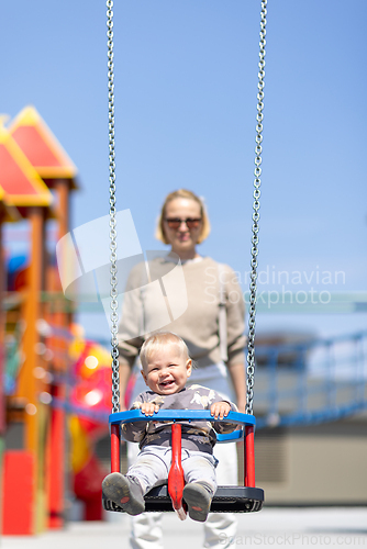Image of Mother pushing her infant baby boy child on a swing on playground outdoors.