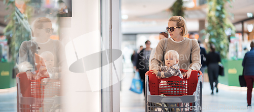 Image of Mother shopping with his kid in the mall. Mother pushing shopping cart with her toddler baby boy child sitting in it along mall corridor.
