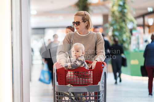 Image of Mother shopping with his kid in the mall. Mother pushing shopping cart with her toddler baby boy child sitting in it along mall corridor.