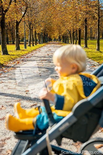 Image of Little toddler child wearing yellow rain coat and rain boots being pushed in stroller by her mother in city park on a sunny autumn day after the rains were gone.