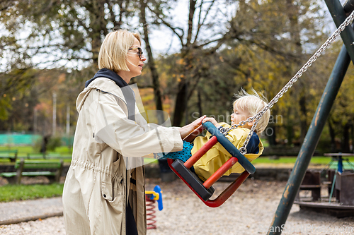 Image of Mother pushing her infant baby boy child wearing yellow rain boots and cape on swing on playground outdoors on cold rainy overcast autumn day in Ljubljana, Slovenia