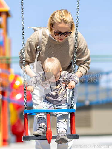 Image of Mother pushing her infant baby boy child on a swing on playground outdoors.