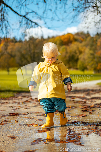 Image of Sun always shines after the rain. Small bond infant boy wearing yellow rubber boots and yellow waterproof raincoat walking in puddles in city park on sunny rainy day.
