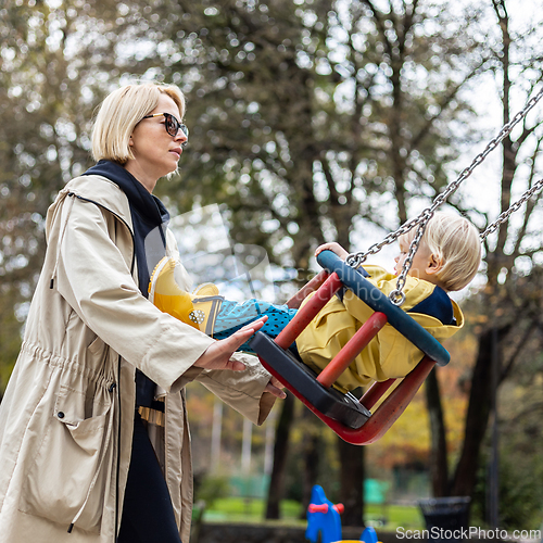 Image of Mother pushing her infant baby boy child wearing yellow rain boots and cape on swing on playground outdoors on cold rainy overcast autumn day in Ljubljana, Slovenia