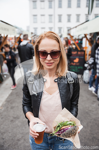 Image of Beautiful young woman holding delicious organic salmon vegetarian burger and homebrewed IPA beer on open air beer an burger urban street food festival in Ljubljana, Slovenia.
