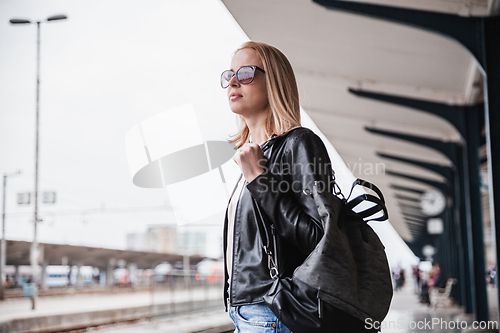 Image of Young blond woman in jeans, shirt and leather jacket wearing bag and sunglass waiting for train to arrive at train station platform. Commuting, travel and transportation concept