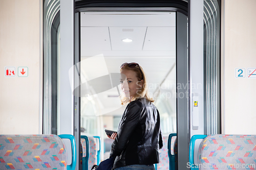 Image of Young blond woman in jeans, shirt and leather jacket holding her smart phone and purse while riding modern speed train arriving to final train station stop. Travel and transportation.
