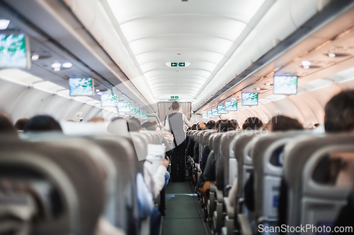 Image of Interior of airplane with passengers on seats and stewardess in uniform walking the aisle, serving people. Commercial economy flight service concept.