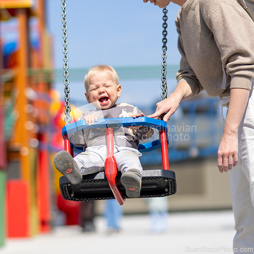 Image of Mother pushing her infant baby boy child on a swing on playground outdoors.