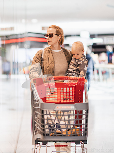 Image of Mother shopping with his kid in the mall. Mother pushing shopping cart while holding and carrying her toddler baby boy child along mall corridor.