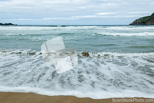 Image of Waves gently lap against the sandy shore under overcast skies at