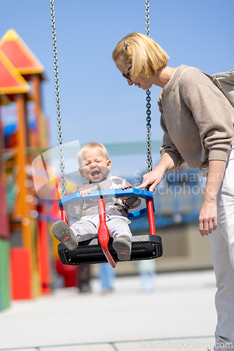 Image of Mother pushing her infant baby boy child on a swing on playground outdoors.