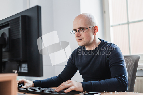 Image of Young Entrepreneur Wearing Eyeglasses Sitting at his Table Inside the Office, Looking at the Report on his Computer Screen.