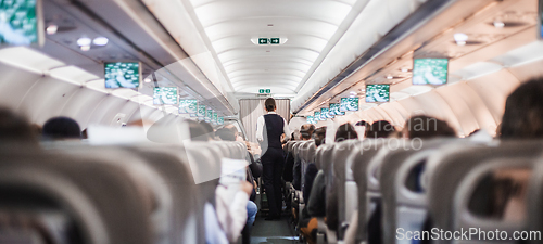 Image of Interior of airplane with passengers on seats and stewardess in uniform walking the aisle, serving people. Commercial economy flight service concept.