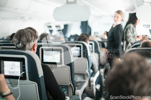 Image of Interior of airplane with passengers on seats and stewardess in uniform walking the aisle, serving people. Commercial economy flight service concept.