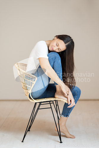 Image of Portrait of confident beautiful woman with long brown hair, wearing casual clothes, sitting on chair in tight jeans and white t-shirt, studio background