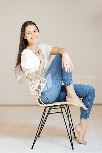 Image of Portrait of confident beautiful woman with long brown hair, wearing casual clothes, sitting on chair in tight jeans and white t-shirt, studio background