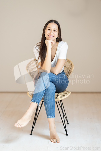 Image of Portrait of confident beautiful woman with long brown hair, wearing casual clothes, sitting on chair in tight jeans and white t-shirt, studio background
