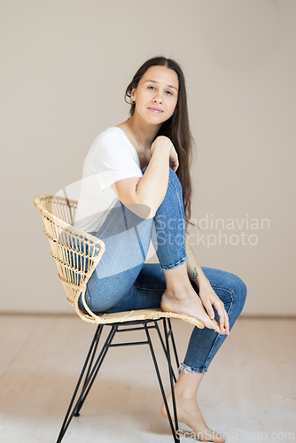 Image of Portrait of confident beautiful woman with long brown hair, wearing casual clothes, sitting on chair in tight jeans and white t-shirt, studio background