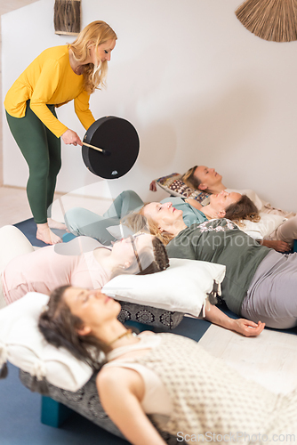 Image of Group therapy session with Tibetan drum for group of women lying on wooden floor surrounded by copper bowls. Meditation and relaxation