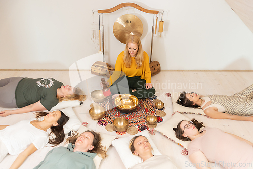Image of Group therapy session with Tibetan singing bowls for group of women lying on wooden floor surrounded by copper bowls. Meditation and relaxation