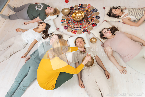 Image of Group therapy session with Tibetan singing bowls for group of women lying on wooden floor surrounded by copper bowls. Meditation and relaxation