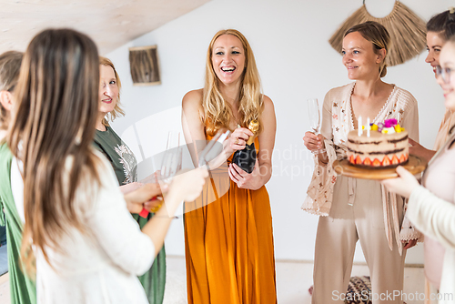 Image of Birthday surprise party. Group of cheerful female friends celebrating personal success stories. Smiling girls toasting with glasses of champagne one holding a birthday cake with candles to blow