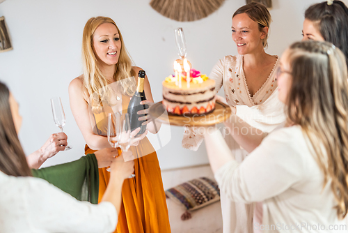Image of Birthday surprise party. Group of cheerful female friends celebrating personal success stories. Smiling girls toasting with glasses of champagne one holding a birthday cake with candles to blow