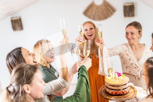 Image of Birthday surprise party. Group of cheerful female friends celebrating personal success stories. Smiling girls toasting with glasses of champagne one holding a birthday cake with candles to blow
