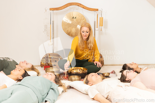Image of Group therapy session with Tibetan singing bowls for group of women lying on wooden floor surrounded by copper bowls. Meditation and relaxation