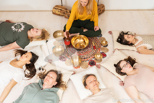 Image of Group therapy session with Tibetan singing bowls for group of women lying on wooden floor surrounded by copper bowls. Meditation and relaxation