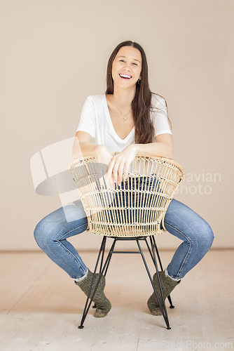 Image of Portrait of confident cheerful beautiful woman with long brown hair, wearing casual clothes, sitting on chair in tight jeans and white t-shirt, studio background