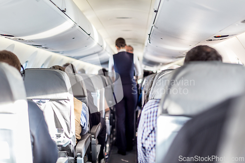 Image of Interior of airplane with passengers on seats and stewardess in uniform walking the aisle, serving people. Commercial economy flight service concept.