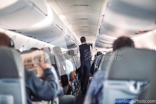 Image of Interior of airplane with passengers on seats and stewardess in uniform walking the aisle, serving people. Commercial economy flight service concept.